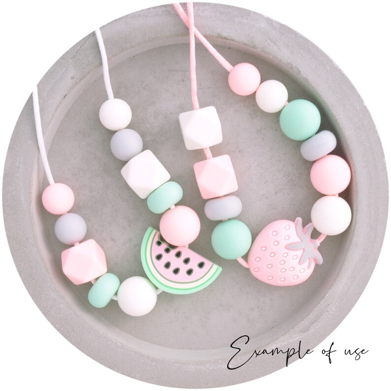 May include: Three silicone teething necklaces with different fruit charms. The necklaces are pink, gray, and white with green accents. The charms are a watermelon, a strawberry, and a square.