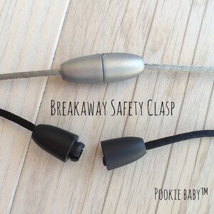 May include: A close-up of a breakaway safety clasp. The image shows a silver clasp connected to a gray wire and a black clasp with a black cord. The text "BREAKAWAY SAFETY CLASP" is printed in black.