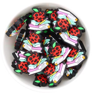 May include: A bowl filled with colourful, red, green and black apple-shaped charms with a stack of books and a pencil. The charms have the words "Teach, Love, Inspire" on them.