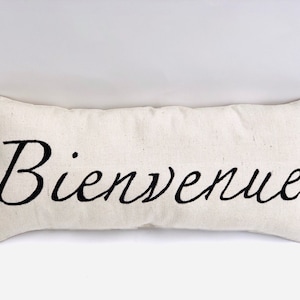 May include: A white rectangular pillow with black embroidered text that reads "Bienvenue".