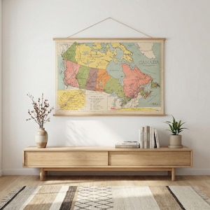 May include: A vintage-style map of Canada, featuring a colorful design with various provinces and territories. The map is displayed on a wooden frame, hanging on a white wall. A wooden cabinet, vase, books, and a plant are below the map.
