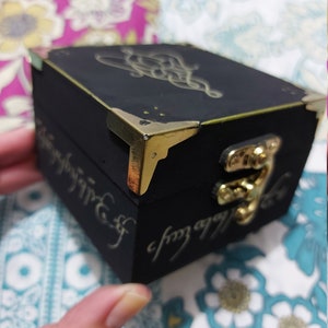 May include: A small black wooden box with gold accents and a gold clasp. The box has a design of a knot on the top and the inscription "One Ring to rule them all" on the side.