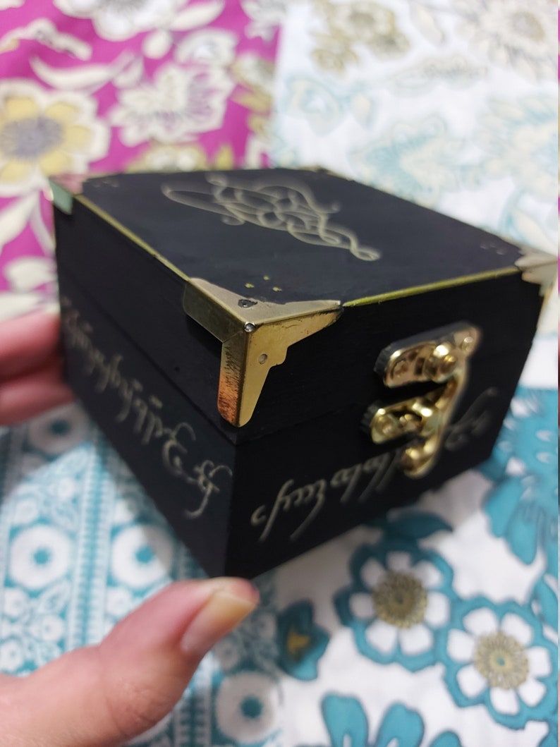 May include: A small black wooden box with gold accents and a gold clasp. The box has a design of a knot on the top and the inscription "One Ring to rule them all" on the side.