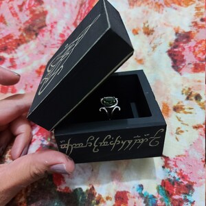 May include: A black wooden ring box with a green gemstone ring inside. The box is engraved with the inscription "One Ring to rule them all, One Ring to find them, One Ring to bring them all and in the darkness bind them."