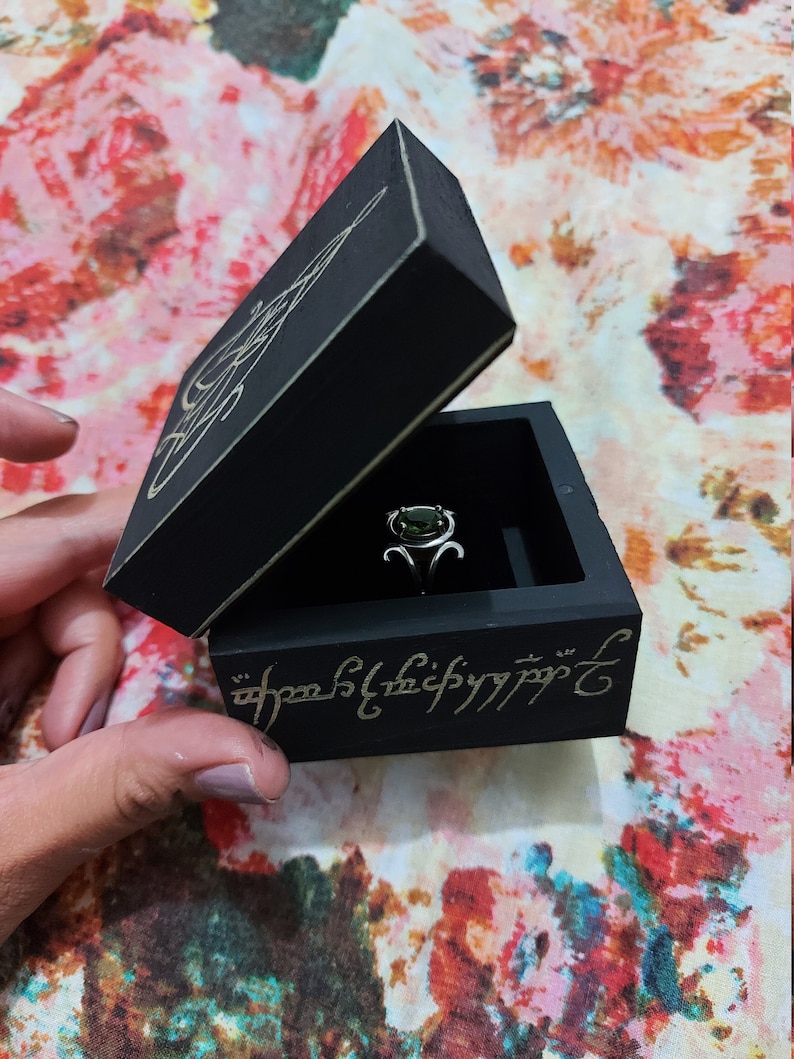May include: A black wooden ring box with a green gemstone ring inside. The box is engraved with the inscription "One Ring to rule them all, One Ring to find them, One Ring to bring them all and in the darkness bind them."