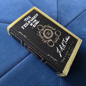 Puede incluir: Un libro de tapa dura titulado "The Fellowship of the Ring" de J.R.R. Tolkien. El libro tiene una cubierta negra con letras e ilustraciones doradas, y bordes dorados. Está sobre una superficie azul. Pertenece a la serie El Señor de los Anillos.