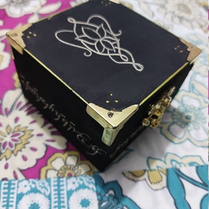 May include: A black wooden box with gold accents and a silver design on the top. The design is a stylized knot with a flower in the centre. The box is sitting on a floral fabric.