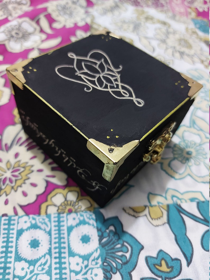 May include: A black wooden box with gold accents and a silver design on the top. The design is a stylized knot with a flower in the centre. The box is sitting on a floral fabric.
