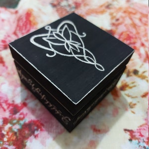 May include: A black wooden box with a white inscription and a white design of a stylised knot. The inscription reads "One Ring to rule them all, One Ring to find them, One Ring to bring them all, and in the darkness bind them."