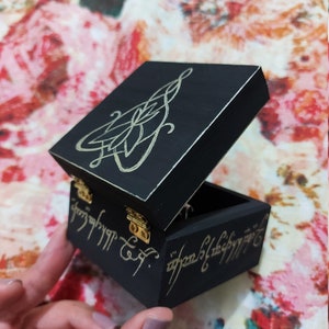May include: A small black wooden box with a gold inscription on the lid and sides. The inscription is in the Elvish language from The Lord of the Rings. The box is open and the inside is black.