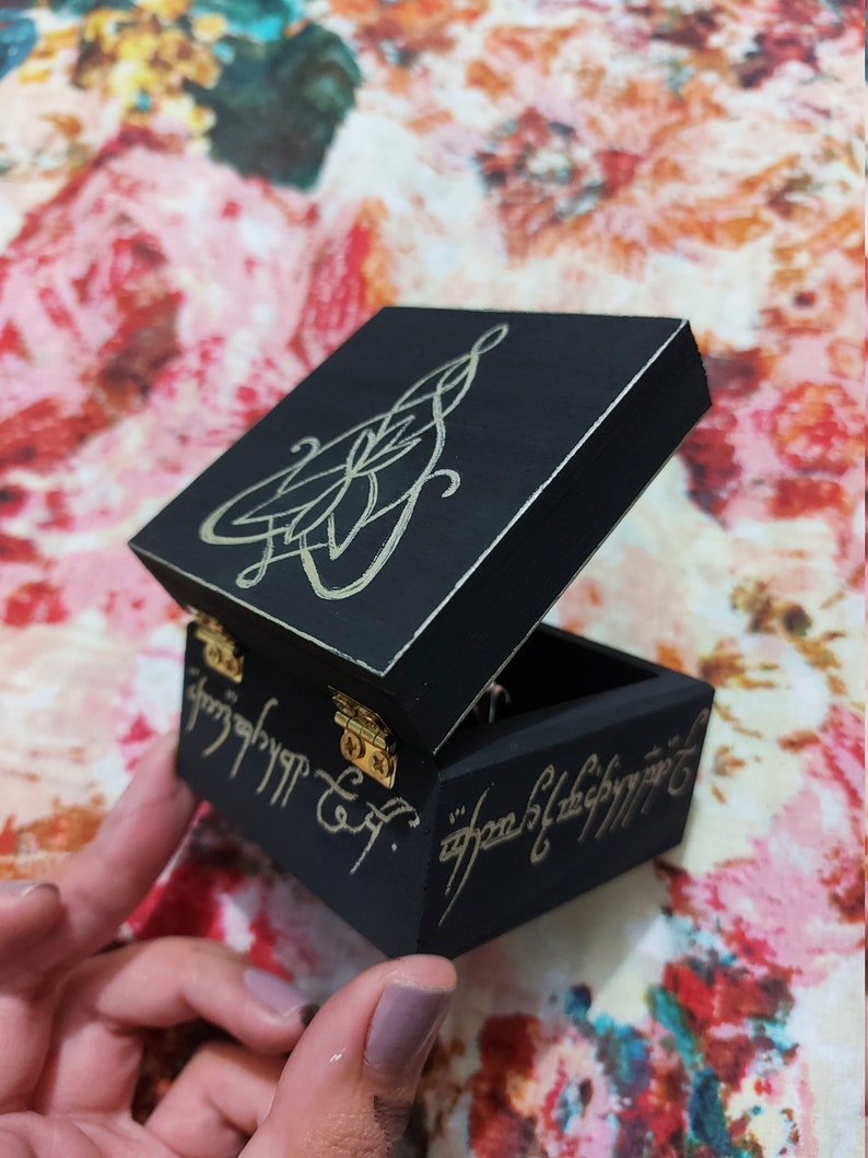 May include: A small black wooden box with a gold inscription on the lid and sides. The inscription is in the Elvish language from The Lord of the Rings. The box is open and the inside is black.
