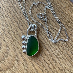 May include: A silver chain necklace with a green sea glass pendant. The pendant is set in silver with three silver beads on the side.