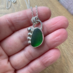 May include: A silver necklace with a green sea glass pendant. The pendant is set in a silver frame with a small silver bead detail.