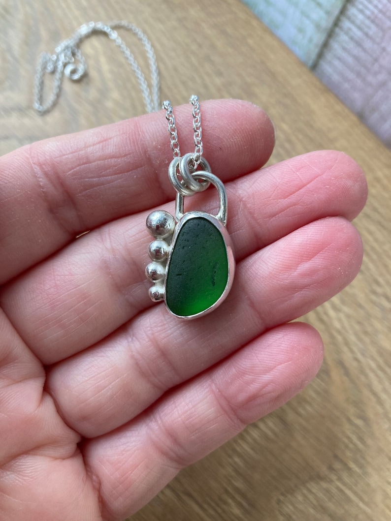 May include: A silver necklace with a green sea glass pendant. The pendant is set in a silver frame with a small silver bead detail.