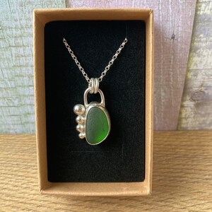 May include: A silver necklace with a green sea glass pendant and silver beads. The necklace is displayed in a brown box with a black velvet lining.