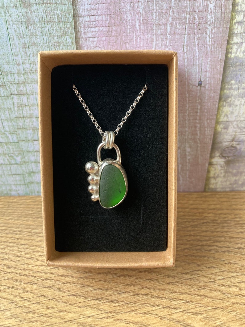May include: A silver necklace with a green sea glass pendant and silver beads. The necklace is displayed in a brown box with a black velvet lining.