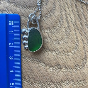 May include: A silver necklace with a green sea glass pendant. The pendant is set in a silver frame with a decorative design. The necklace is approximately 2 inches long.