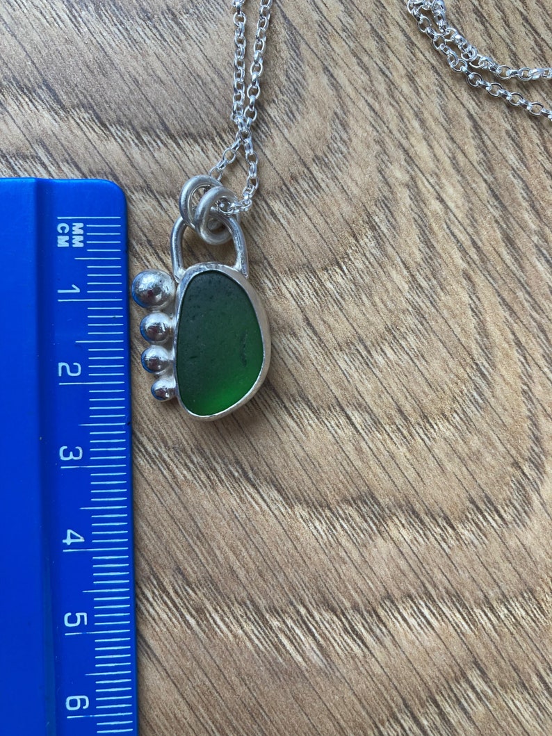 May include: A silver necklace with a green sea glass pendant. The pendant is set in a silver frame with a decorative design. The necklace is approximately 2 inches long.