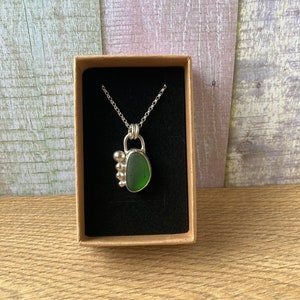 May include: A silver necklace with a green sea glass pendant. The pendant is set in a silver bezel and has a cluster of silver beads on the side.