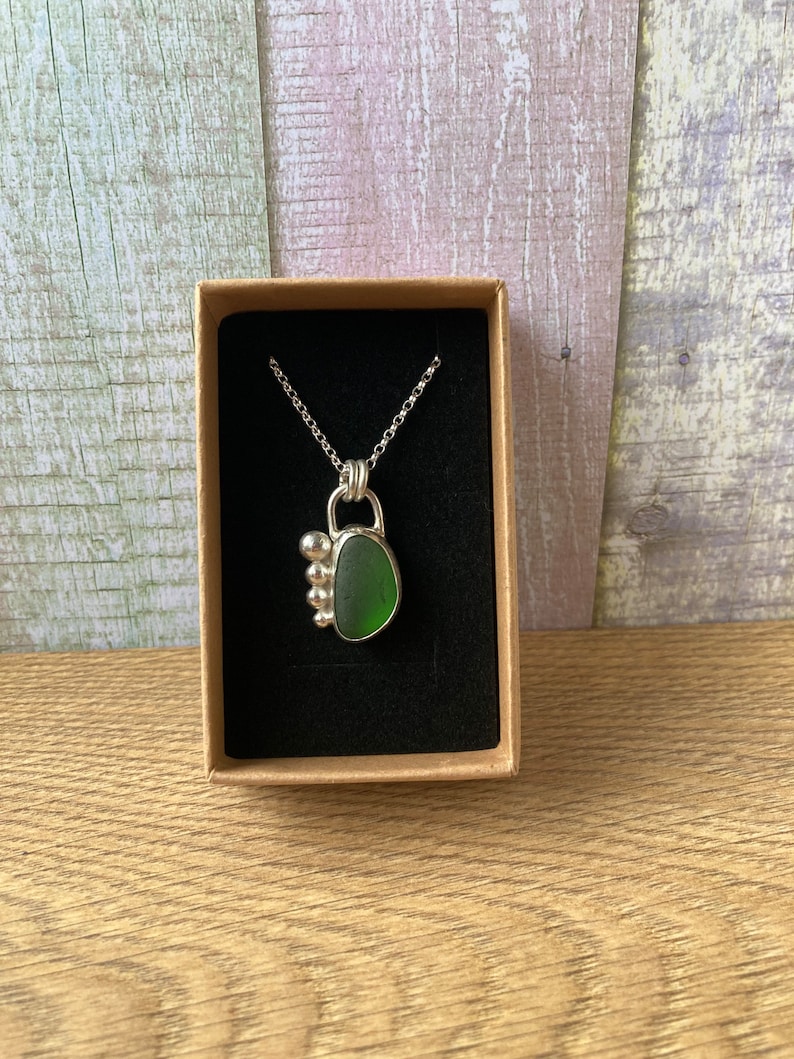 May include: A silver necklace with a green sea glass pendant. The pendant is set in a silver bezel and has a cluster of silver beads on the side.