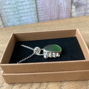 May include: A silver necklace with a green sea glass pendant. The pendant is set in a silver frame with a decorative design. The necklace is displayed in a brown box with a black interior.