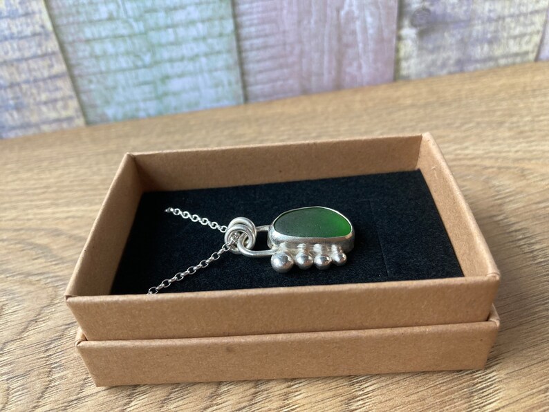 May include: A silver necklace with a green sea glass pendant. The pendant is set in a silver frame with a decorative design. The necklace is displayed in a brown box with a black interior.