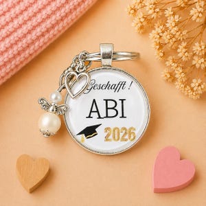 May include: A silver keychain with a round pendant featuring the text "Geschafft! ABI 2026" and a graduation cap graphic. The keychain includes a heart charm, an angel charm with a pearl, and a key ring. The background is peach-colored.