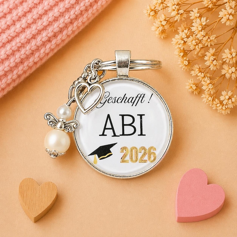 May include: A silver keychain with a round pendant featuring the text "Geschafft! ABI 2026" and a graduation cap graphic. The keychain includes a heart charm, an angel charm with a pearl, and a key ring. The background is peach-colored.