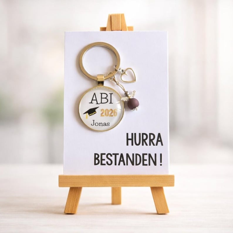 Pu&ograve; includere: Un portachiavi a tema laurea con anello e ciondoli color oro. Il ciondolo rotondo mostra "ABI 2026" con un tocco e il nome "Jonas". La carta recita "HURRA BESTANDEN!" su un piccolo cavalletto di legno.