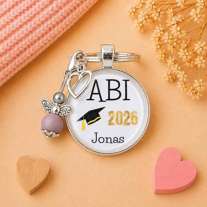 Pu&ograve; includere: Un portachiavi argentato con un ciondolo rotondo con la scritta "ABI 2026 Jonas" e una grafica di cappello di laurea. Il portachiavi include un ciondolo a forma di angelo con una perla viola e un ciondolo a forma di cuore.