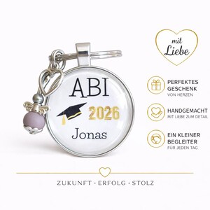 Pu&ograve; includere: Un portachiavi argentato con un ciondolo rotondo con la scritta "ABI 2026 Jonas" e una grafica di cappello di laurea. Il portachiavi include un ciondolo decorativo con un cuore e una perla lavanda. L'immagine include anche testo in tedesco.