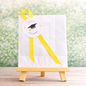 May include: A white paper gift bag with a yellow ribbon and a circular label. The label reads "ABITUR 2026" and features a graduation cap illustration. The bag is displayed on a small wooden easel.