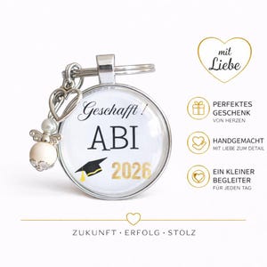 May include: A silver keychain with a round pendant featuring the text "Geschafft! ABI 2026" and a graduation cap graphic. The keychain includes a heart charm and an angel-shaped bead. The image also includes text in German.