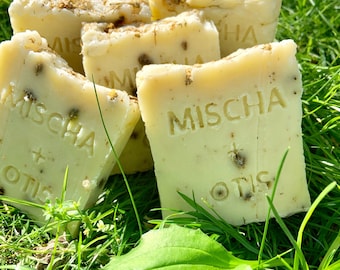 May include: Four bars of handmade soap with the words "Mischia + Otis" printed on them. The soap is a light yellow color and is sitting on a bed of green grass.