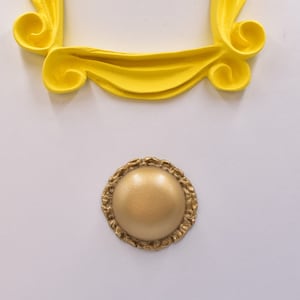 Puede incluir: Un marco de plástico amarillo en forma de marco de imagen con un botón redondo dorado en el centro.