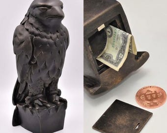 Könnte beinhalten: Eine dunkel bronzefarbene Falkenfigur mit einem hohlen Innenraum, der als Spardose dient. Die Skulptur verfügt über eine abnehmbare quadratische Bodenplatte und ist mit einem in den Schlitz gesteckten Dollarschein sowie einer kupferfarbenen Bitcoin-Münze abgebildet.