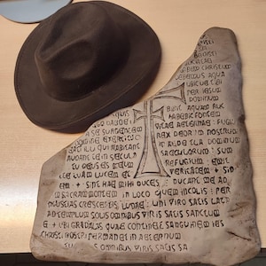 Könnte beinhalten: Ein brauner Fedora-Hut liegt neben einer verwitterten, cremefarbenen Steintafel. Die Tafel zeigt ein eingeritztes Kreuz und lateinischen Text. Der Hut hat eine klassische Tränenform und eine breite Krempe. Die Gegenstände liegen auf einer hellbraunen Oberfläche.