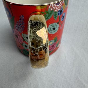 May include: A red ceramic mug with a floral pattern in pink, blue, and white. The handle is clear with gold accents and contains small decorative elements. The rim of the mug has a gold trim.