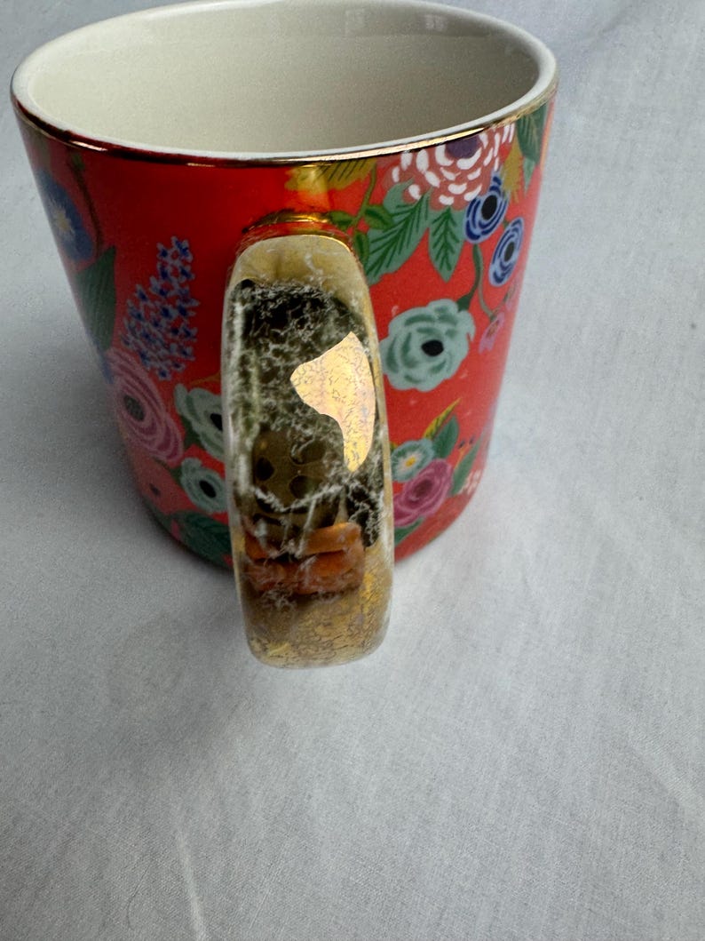 May include: A red ceramic mug with a floral pattern in pink, blue, and white. The handle is clear with gold accents and contains small decorative elements. The rim of the mug has a gold trim.