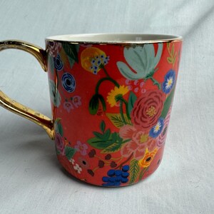 May include: A ceramic mug with a vibrant floral pattern in red, pink, blue, and yellow. The mug has a gold-colored handle and rim, adding a touch of elegance. The design features various flowers and foliage.