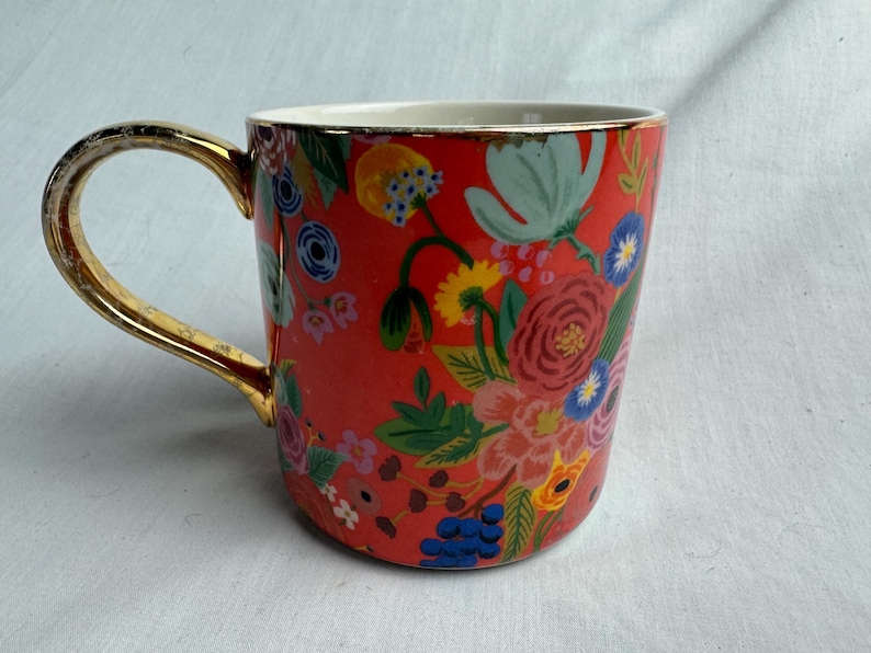 May include: A ceramic mug with a vibrant floral pattern in red, pink, blue, and yellow. The mug has a gold-colored handle and rim, adding a touch of elegance. The design features various flowers and foliage.