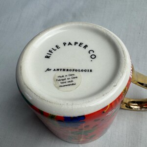 May include: A ceramic mug with a colorful floral design and a gold handle. The bottom of the mug features the text "RIFLE PAPER CO." and "for ANTHROPOLOGIE". A small sticker indicates the mug is made in China and hand wash is recommended.