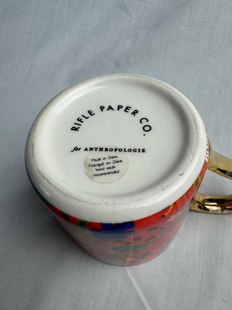 May include: A ceramic mug with a colorful floral design and a gold handle. The bottom of the mug features the text "RIFLE PAPER CO." and "for ANTHROPOLOGIE". A small sticker indicates the mug is made in China and hand wash is recommended.