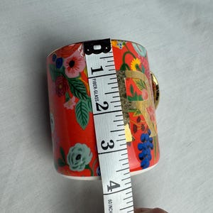 May include: A red floral ceramic mug with a gold handle and the letter "R". The mug is approximately 4 inches tall. The design includes various colorful flowers and foliage.