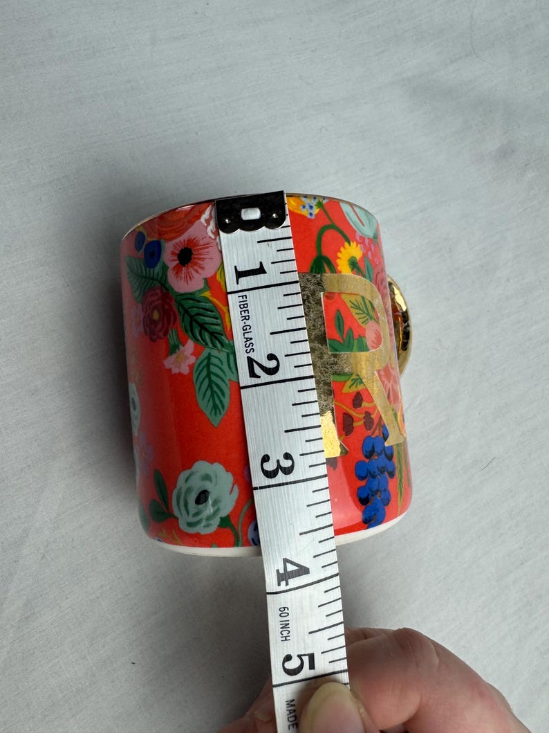 May include: A red floral ceramic mug with a gold handle and the letter "R". The mug is approximately 4 inches tall. The design includes various colorful flowers and foliage.