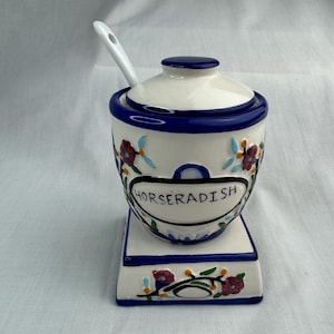 Puede incluir: Tarro de cerámica blanca para rábano picante con borde y tapa azules, diseños florales y la palabra "HORSERADISH" en un óvalo negro. Incluye una pequeña cuchara blanca y se asienta sobre una base rectangular a juego.