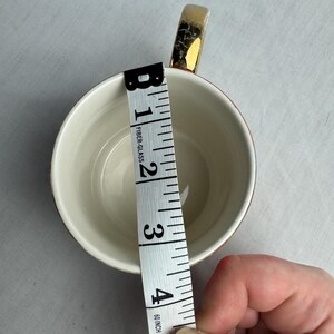 May include: A white teacup with a gold handle and rim, measuring approximately 4 inches in diameter. A measuring tape is placed inside the cup to show the diameter. The cup is on a white surface.