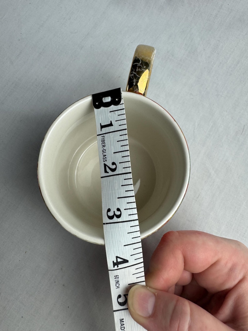 May include: A white teacup with a gold handle and rim, measuring approximately 4 inches in diameter. A measuring tape is placed inside the cup to show the diameter. The cup is on a white surface.