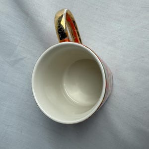 May include: A white ceramic mug with a gold-colored handle and rim. The mug has a red and black patterned design on the exterior. The mug is empty and sits on a light gray surface.