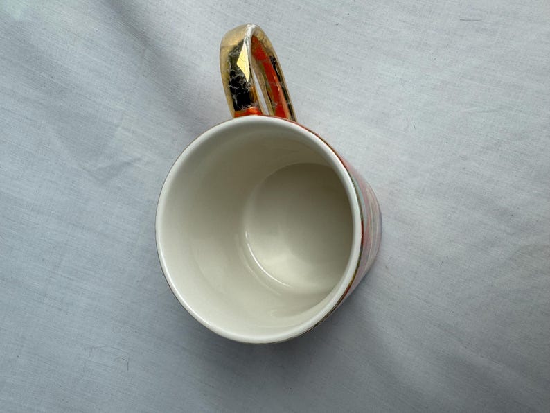 May include: A white ceramic mug with a gold-colored handle and rim. The mug has a red and black patterned design on the exterior. The mug is empty and sits on a light gray surface.